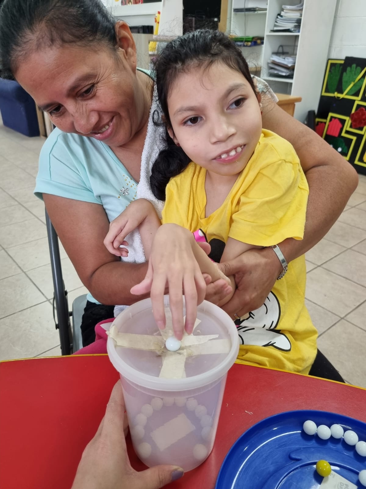 Dos personas participando en una actividad de motricidad fina, utilizando objetos pequeños y un contenedor para desarrollar la precisión y la coordinación de los movimientos de los dedos.